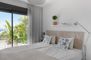 A bedroom with a large bed, a view of greenery and buildings, a shelf with a plant and a shell, and a bedside lamp.