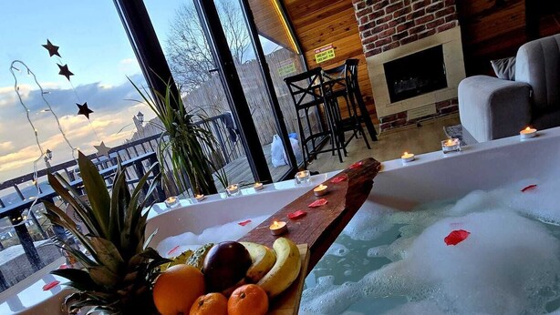 Un espace de détente avec un jacuzzi, une table en bois agrémentée de fruits et de bougies, et une vue sur le paysage urbain grâce à de grandes fenêtres.