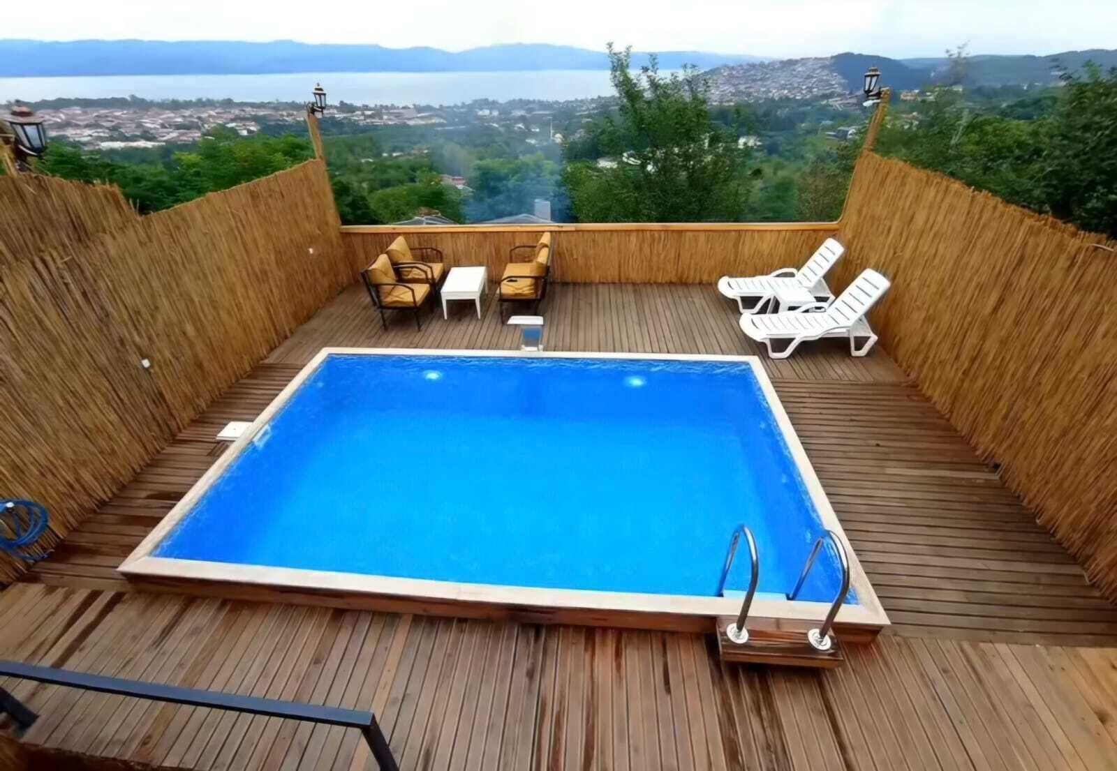 Una piscina rettangolare con un prendisole in legno, circondata da poltroncine e una recinzione in legno.