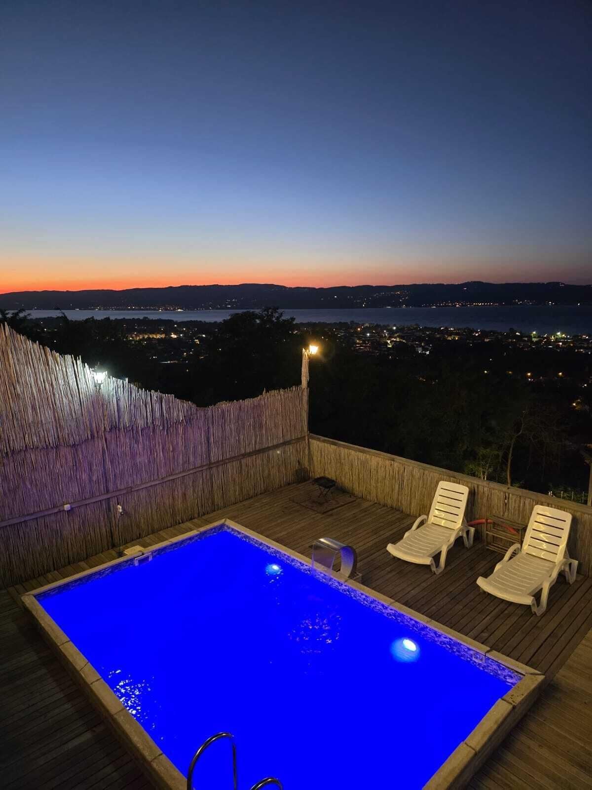 Piscina externa retangular com um deck de madeira, duas espreguiçadeiras e vista para um horizonte urbano ao entardecer.