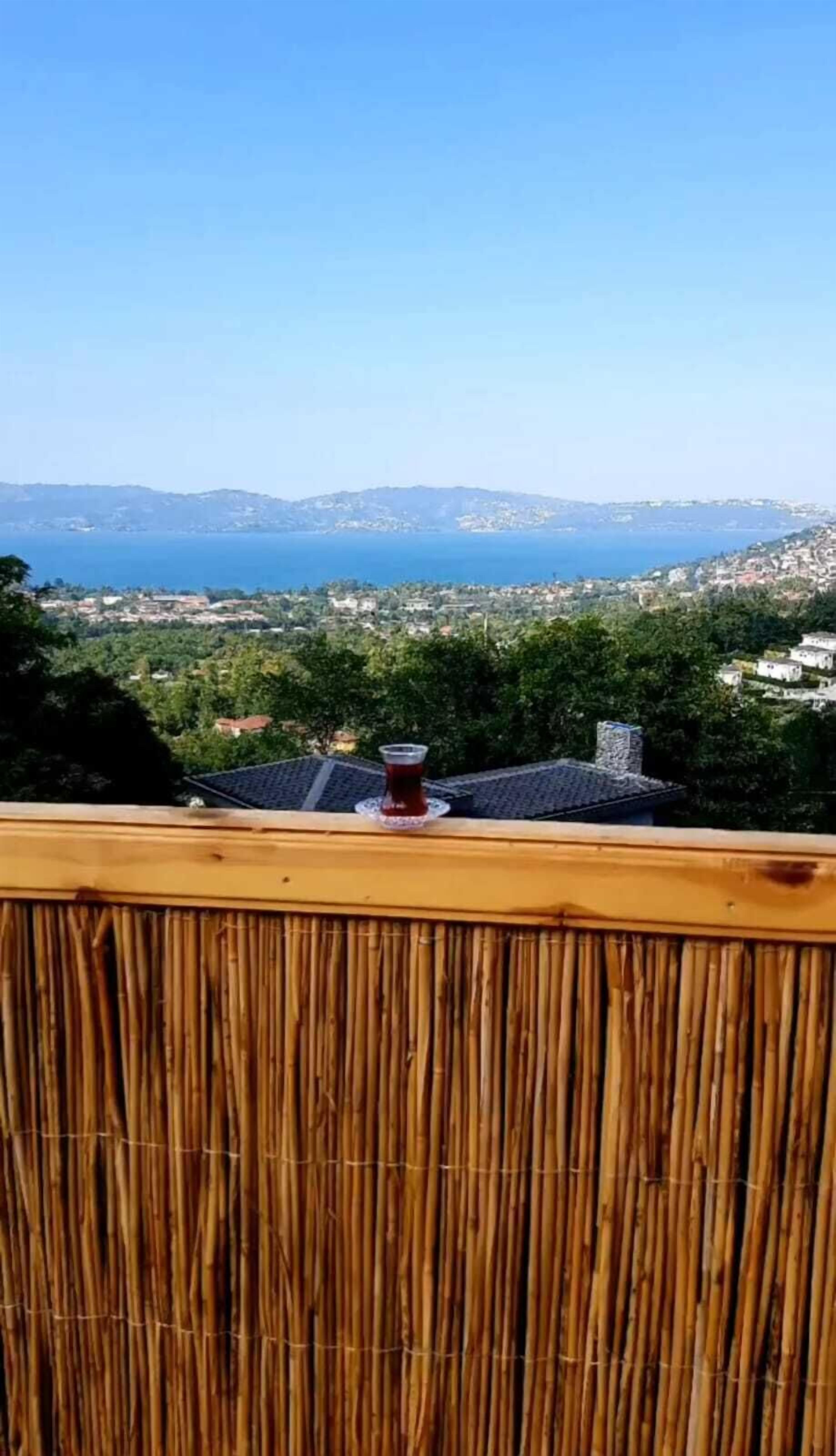 Une vue depuis un balcon, avec une clôture en bambou, un verre contenant un liquide rouge et une vue lointaine sur la ville.