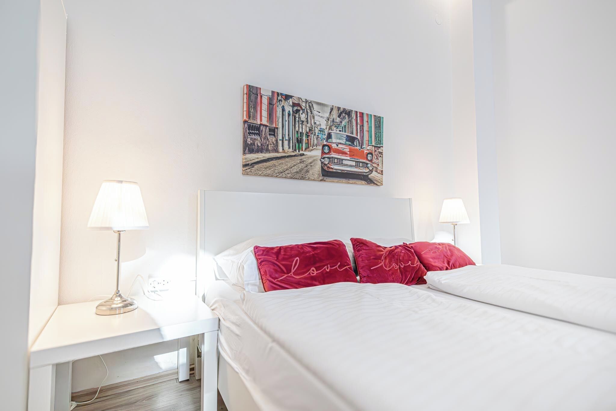 A hotel room with a bed, two bedside lamps, a white nightstand, and a colorful painting above the bed.