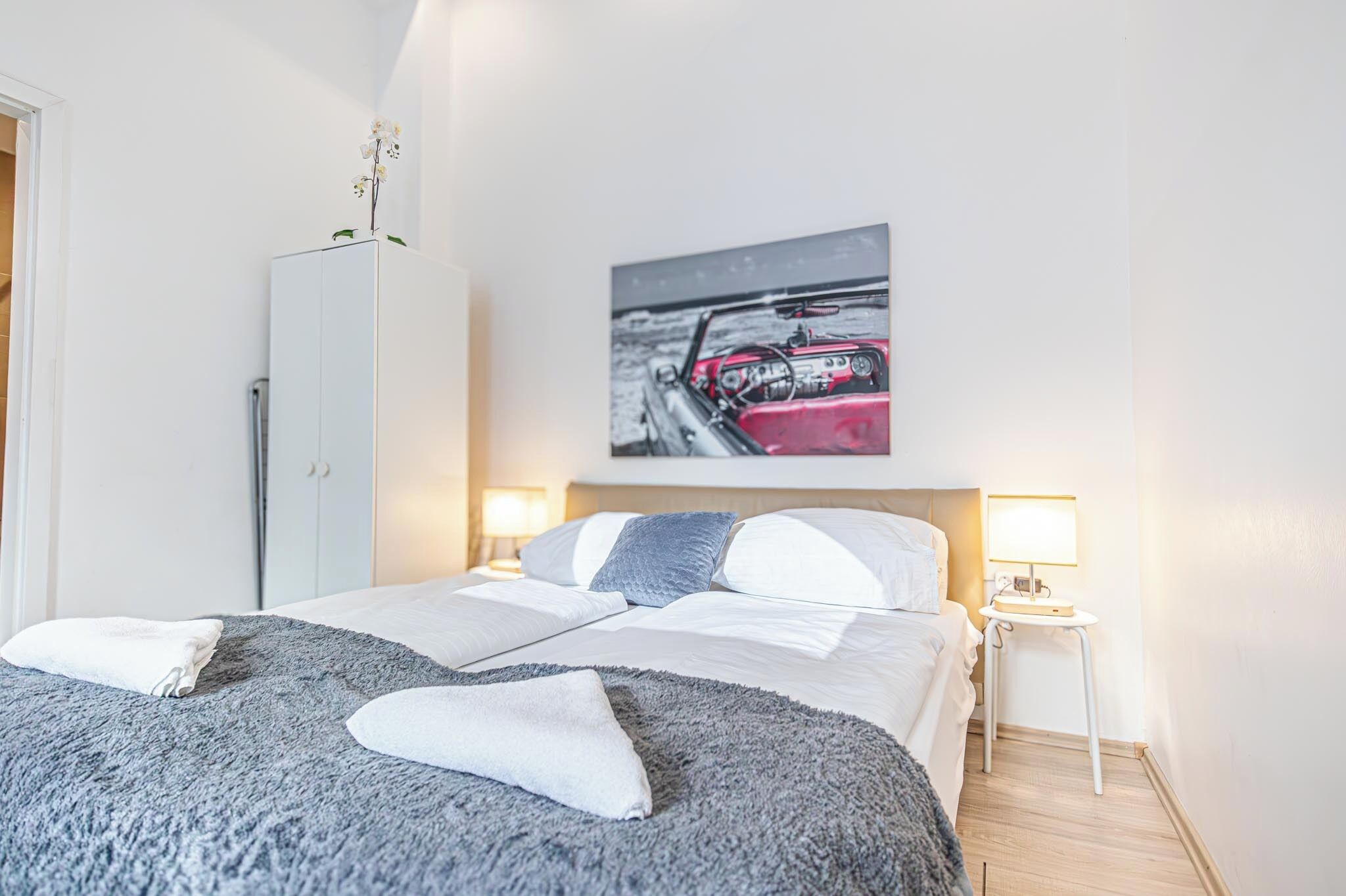A neatly made bed with a grey blanket and pillows, a white wardrobe, a bedside table with a lamp, and a wall-mounted picture of a red car.