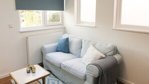 Una habitación con un sofá azul claro, una mesita con objetos decorativos y dos ventanas con marcos blancos.
