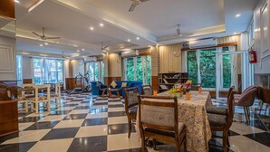 A spacious hotel lobby with a checkered floor, multiple seating areas, ceiling fans, and large windows offering views of greenery. - Alaya Stays Impreza Luxury stays (Dehradun)
