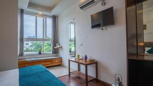 A modern hotel room with a large window, a bed with a teal bedspread, a wooden bench, a small table with books and a lantern, and a mounted TV. - Alaya Stays Impreza Luxury stays (Dehradun)