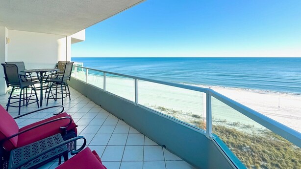 A balcony with a view of the ocean, a table with chairs, and a red lounge chair. - Silver Beach 806 | Top Floor, Corner, Beach Front - Penthouse Upgrades! (Orange Beach)