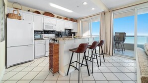 A modern kitchen with white cabinets, a microwave, and a stove. There are three bar stools and a wicker basket. The kitchen has a large window with a view of the ocean. - Silver Beach 806 | Top Floor, Corner, Beach Front - Penthouse Upgrades! (Orange Beach)