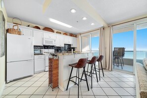 A modern kitchen with white cabinets, a microwave, and a stove. There are three bar stools and a wicker basket. The kitchen has a large window with a view of the ocean. - Silver Beach 806 | Top Floor, Corner, Beach Front - Penthouse Upgrades! (Orange Beach)