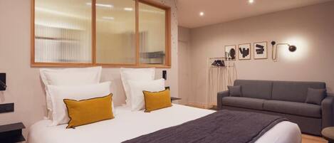 A modern hotel room with a large bed, a grey sofa, and a window with blinds.
