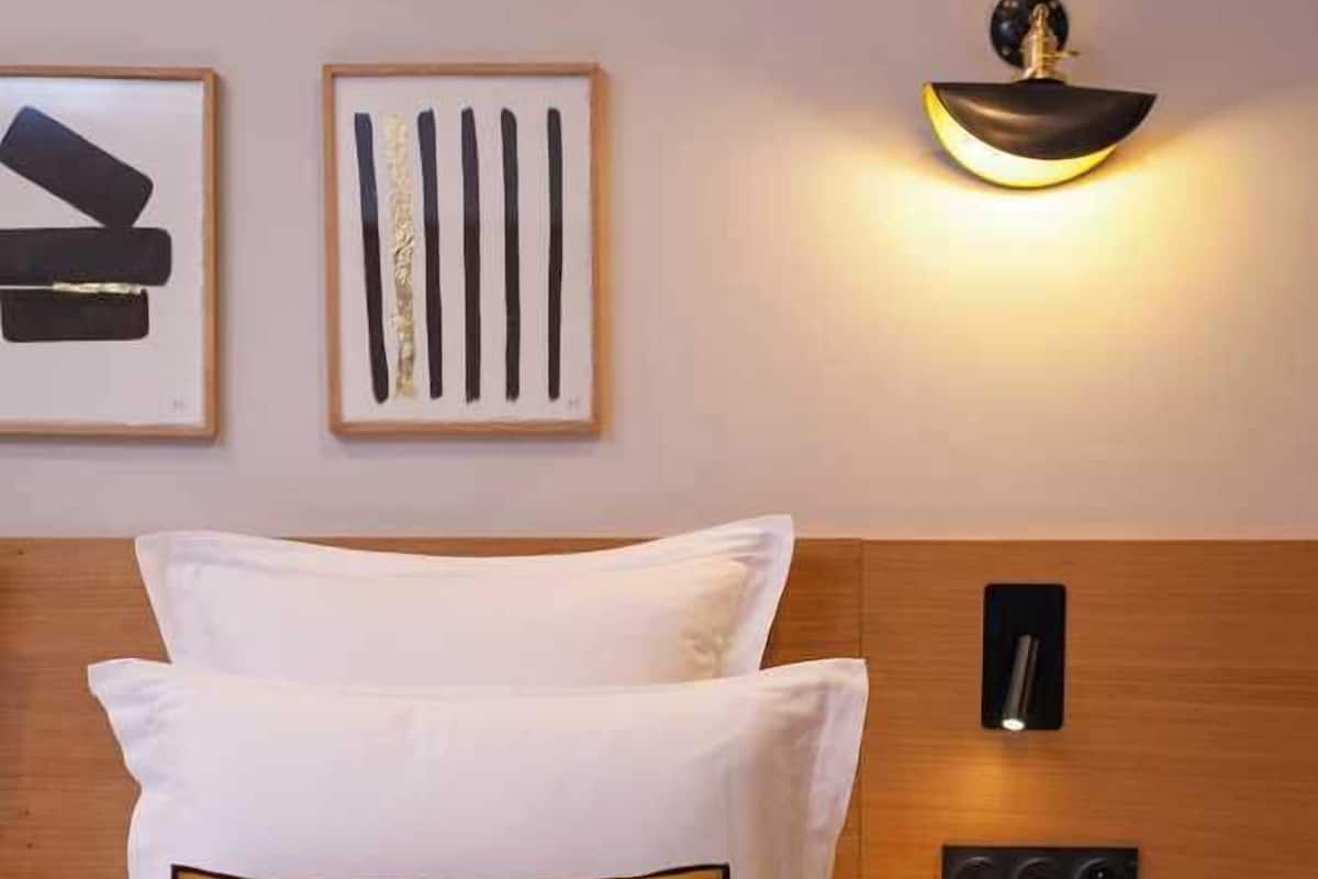 A neatly made bed with white and mustard pillows, a wooden headboard, a wall-mounted light fixture, and a small bedside table with a remote control.