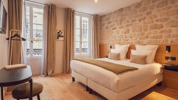 Une chambre avec un grand lit, une petite table et une chaise. On y trouve une fenêtre avec des rideaux, un mur en pierre et un balcon visible à travers la fenêtre.