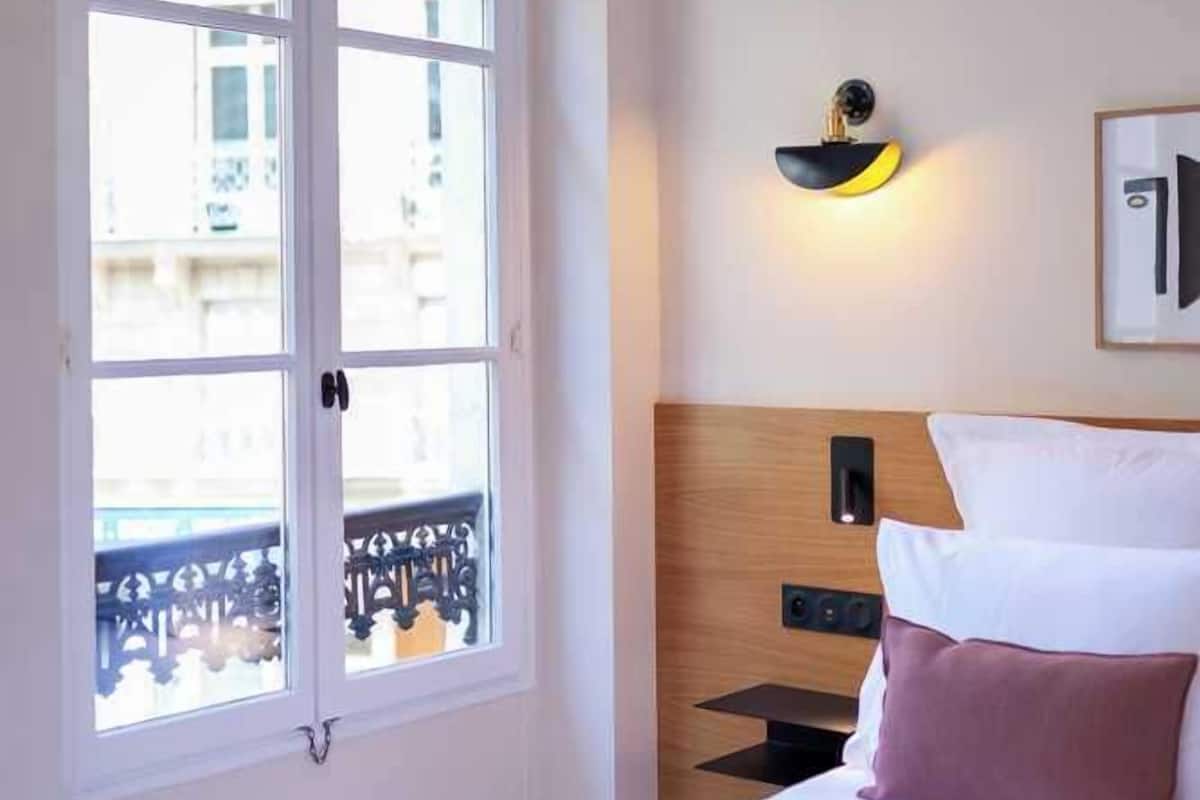 A hotel room with a bed, a nightstand, a wall-mounted lamp, and a window with a view of buildings.