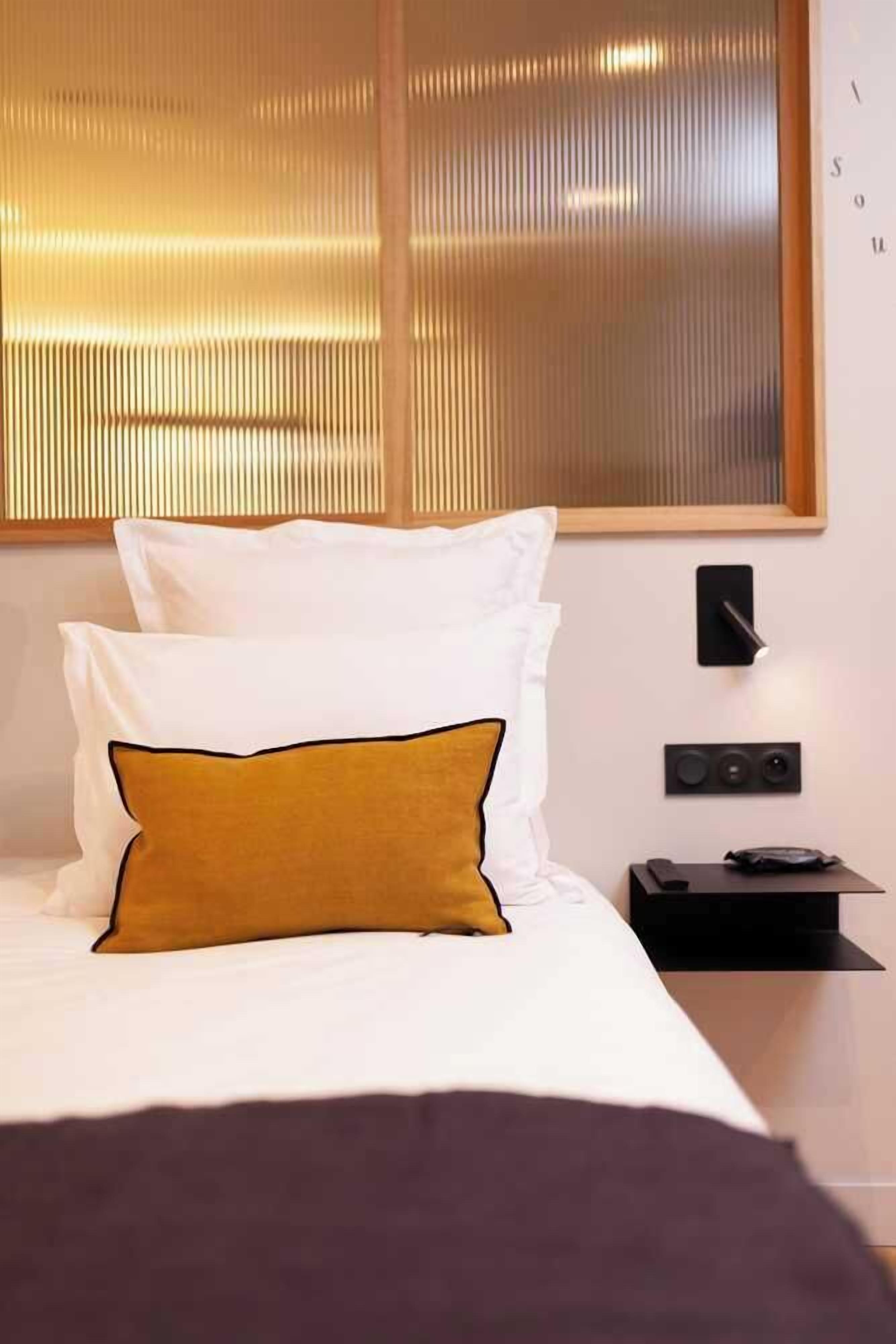 A neatly made bed with white and mustard pillows, a black bedside table with a phone and a small speaker, and a wall-mounted light.