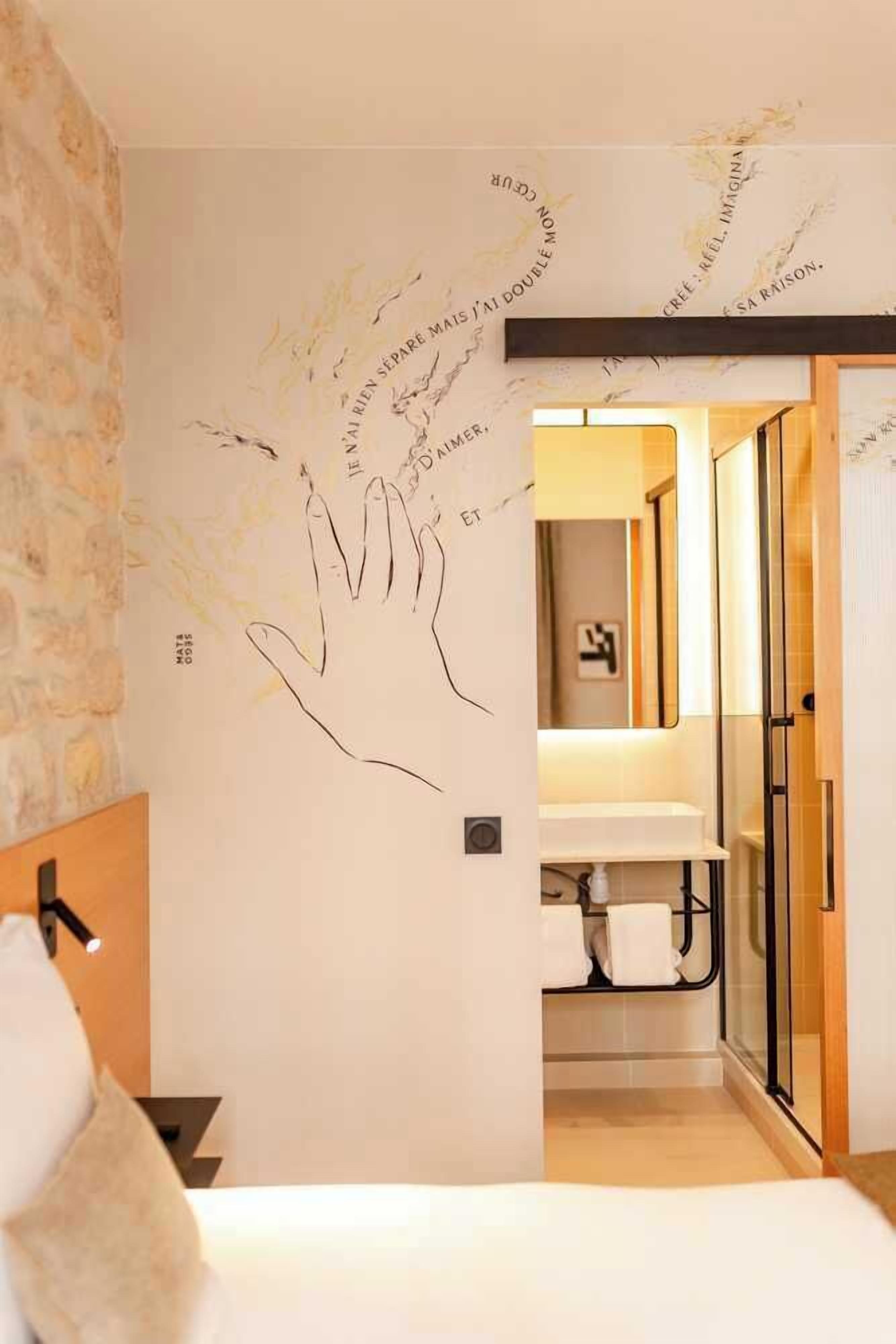 Una camera d'albergo con un grande disegno a mano sulla parete, un letto, una testiera in legno, un comodino e un bagno visibile attraverso una porta aperta.