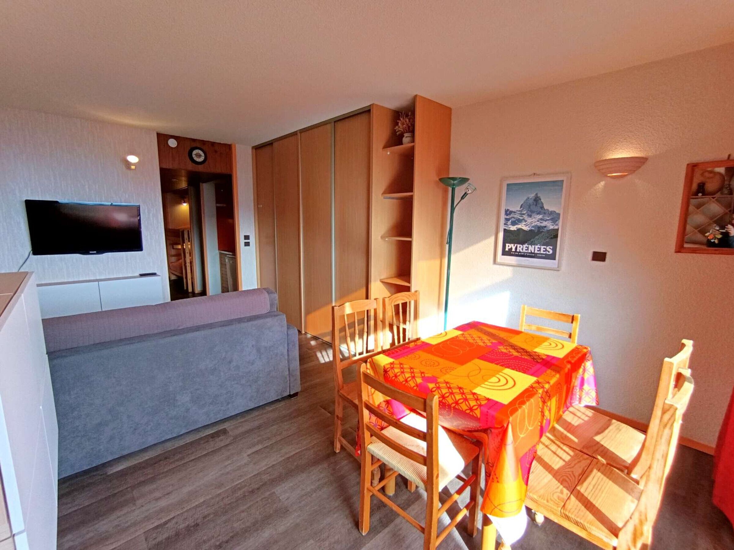 A compact living space with a sofa, a dining table with chairs, a TV mounted on the wall, and a poster of the Pyrenees.