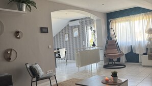 Un saló modern amb un sofà gris, una taula de centre amb una espelma i una cadira penjant. Es pot veure una zona de menjador i una cuina al fons.