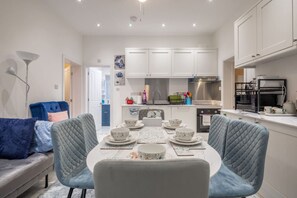 Eine moderne Küche mit weißen Schränken, ein Essbereich mit einem runden Tisch und blauen Stühlen sowie eine Wohnarea mit einer Couch und einem blauen Sessel.