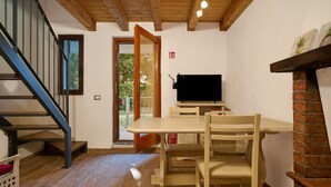 Een kamer met een houten trap, een televisie aan een bakstenen muur, een eettafel met stoelen en een schuifdeur die naar buiten leidt.
