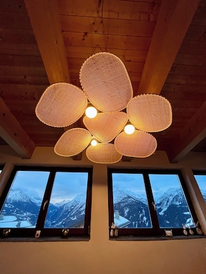 A room with a wooden ceiling and a woven light fixture. There are three windows with a view of snow-covered mountains.