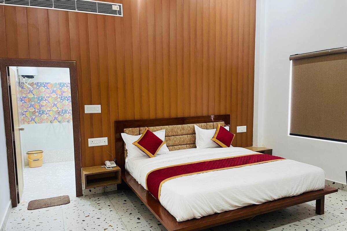 Habitación de hotel con cabecera de madera, cama con ropa blanca y detalles rojos, mesita de noche con un teléfono y baño con zona de ducha colorida.