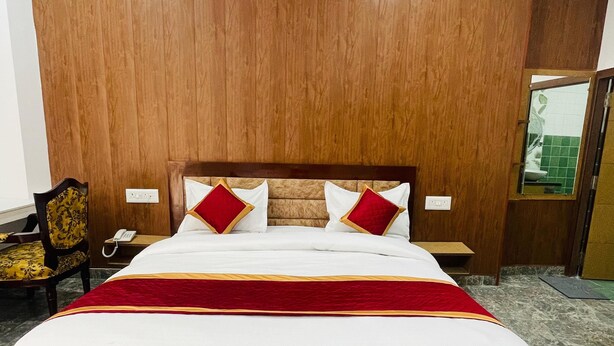 Una cama grande con cabecera de madera, ropa de cama blanca y detalles en rojo. Hay una silla con asiento estampado y una mesita con un teléfono. La habitación tiene piso de mármol y una puerta de madera con una ventana.