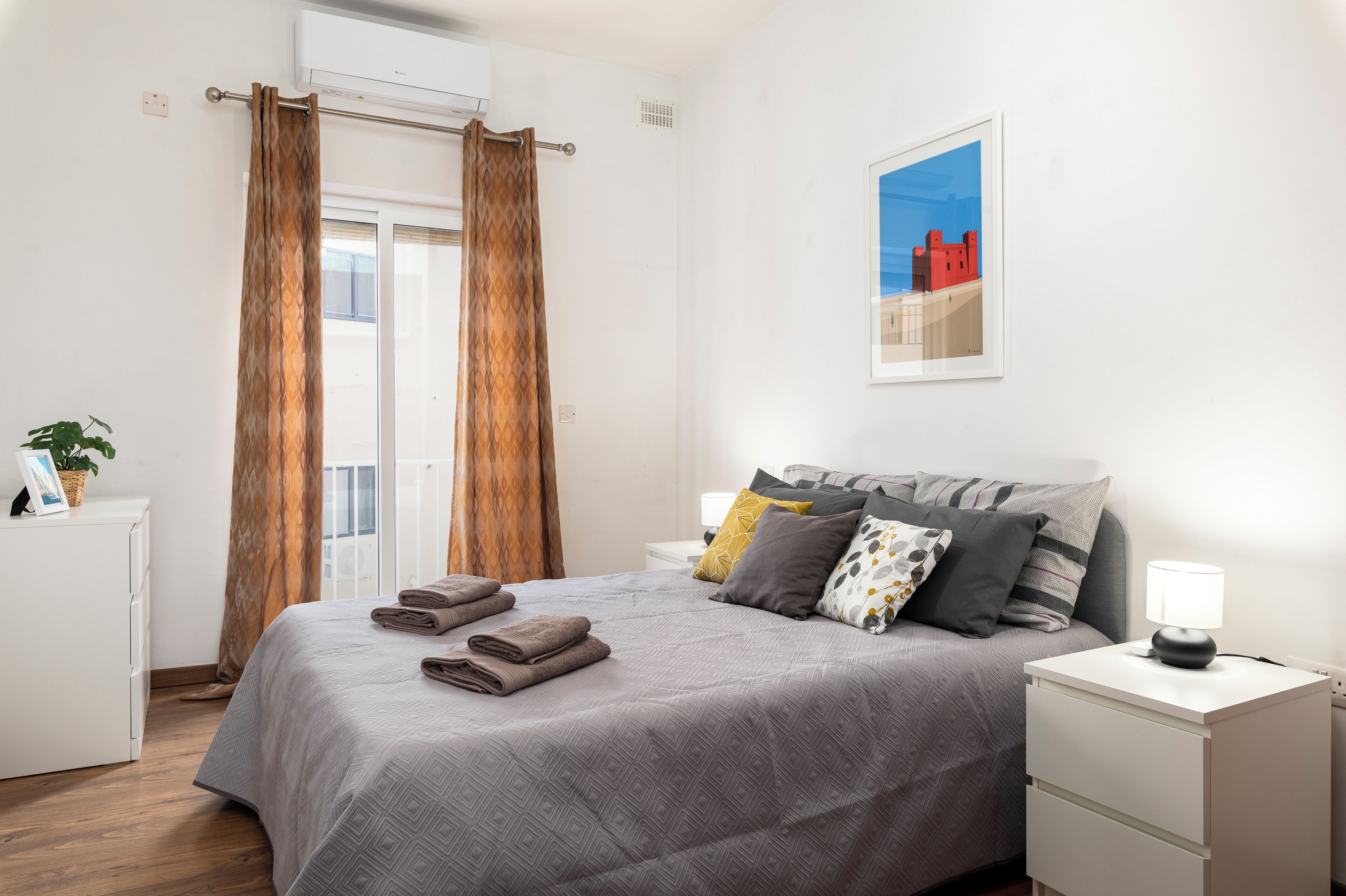 A neatly made bed with a grey patterned bedspread, a bedside lamp, and a framed picture on the wall.
