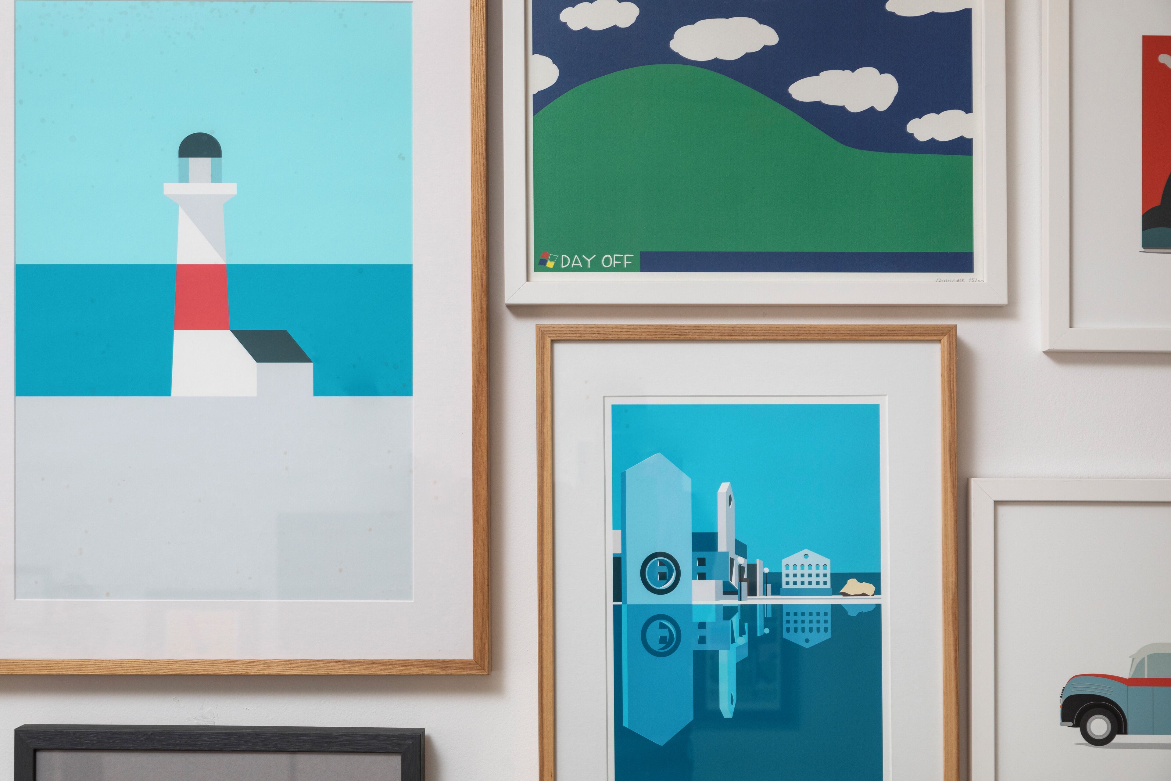A wall with multiple framed artworks, including a lighthouse, a green hill with clouds, and a cityscape with a bridge.