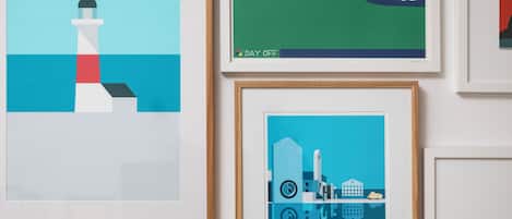 A wall with multiple framed artworks, including a lighthouse, a green hill with clouds, and a cityscape with a bridge.