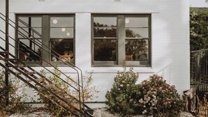 A white building with a staircase leading to a door, a window with a view of a person, and a small garden with flowers. - The Kentish Tasmania (Oatlands)