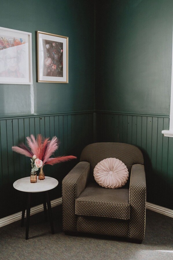 A corner room with a green wall, a patterned armchair with a pink pillow, a small round table with a vase of pink flowers, and two framed pictures on the wall.