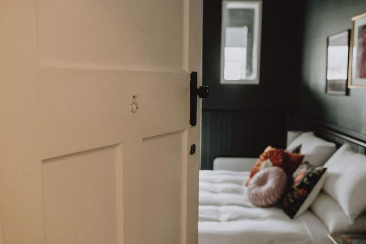 A hotel room with a white door, a bed with white bedding, and a dark wall with a window and framed pictures.
