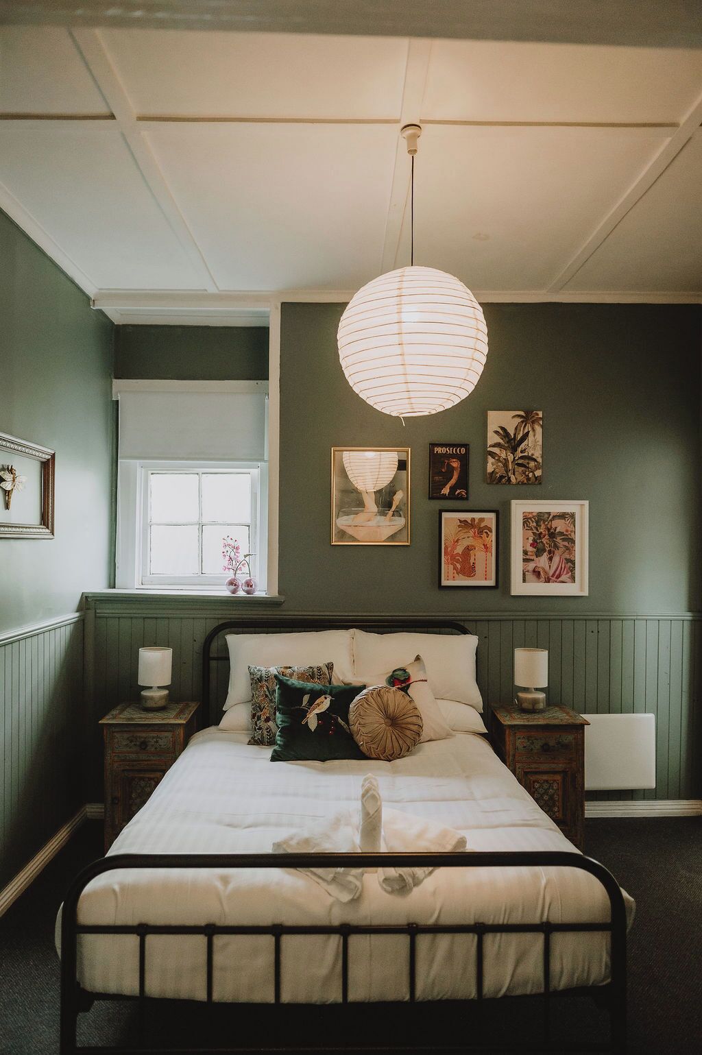 A bedroom with a bed, bedside tables, a hanging light fixture, and wall art.