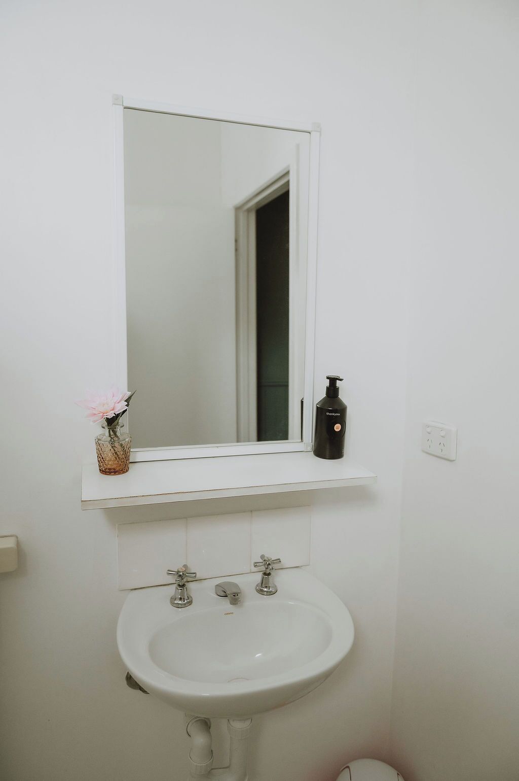 Un baño con un lavabo blanco, un espejo encima y un estante pequeño con una botella y un florero.