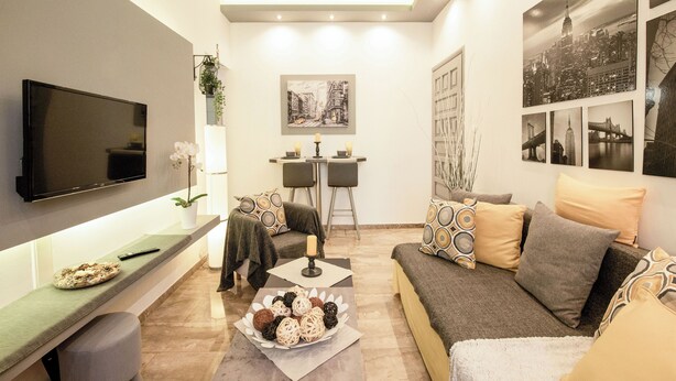 Ein modernes Wohnzimmer mit einem Flachbildfernseher, einer Couch, einem Couchtisch mit Dekoration und einem Essbereich mit zwei Stühlen und einem kleinen Tisch.
