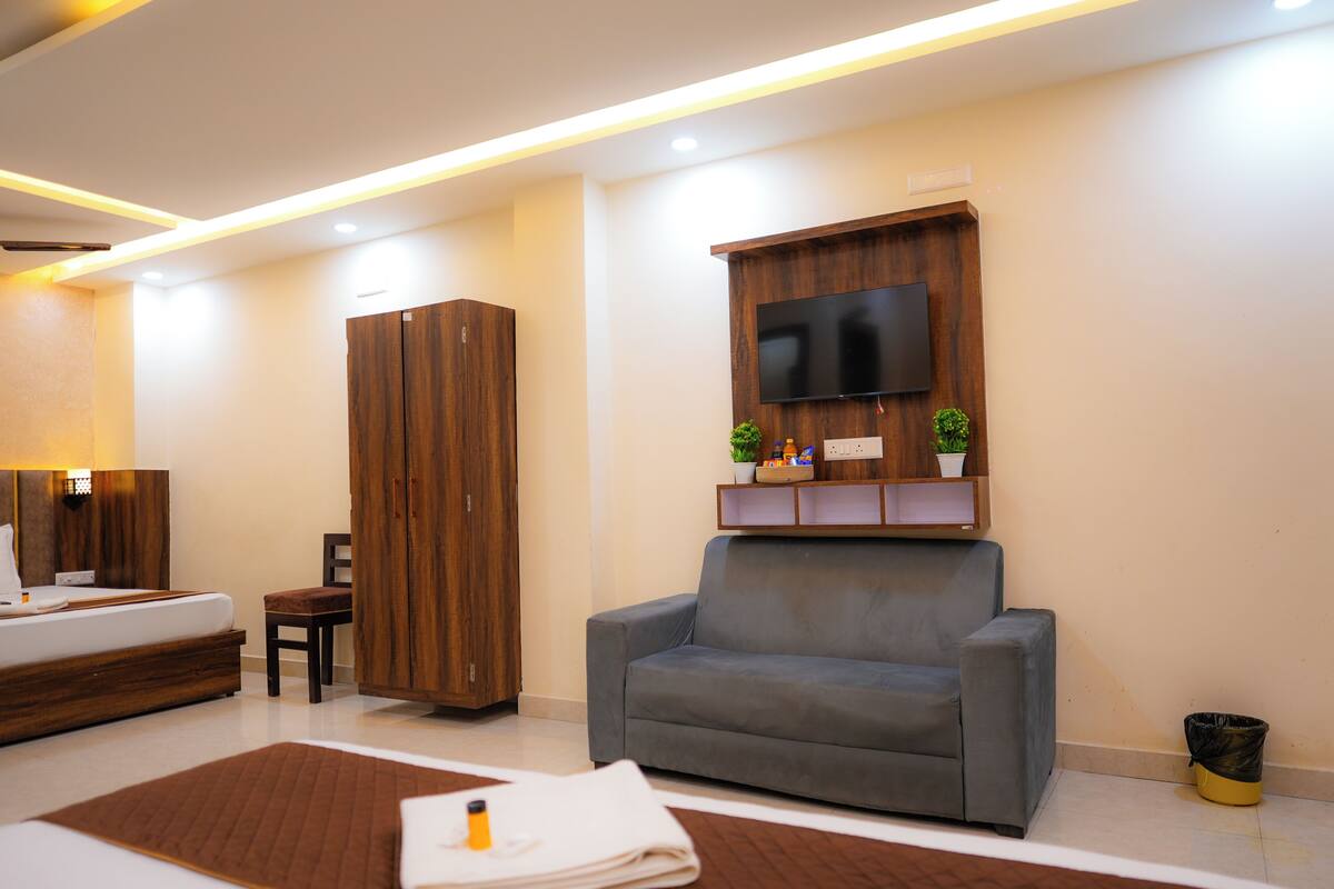 A modern hotel room with a bed, a grey sofa, a wooden wardrobe, a TV mounted on the wall, and a small shelf with plants and toiletries.