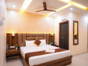 Une chambre d’hôtel moderne dotée d’un grand lit, d’une tête de lit en bois, de tables de chevet, d’une petite table, d’un ventilateur de plafond, d’un miroir fixé au mur et d’une porte au design original.