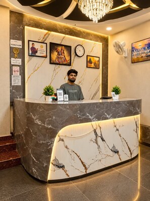 A hotel reception desk with a marble design, framed artwork, a clock, and a chandelier. - Shree Hari Kunj (Varanasi)