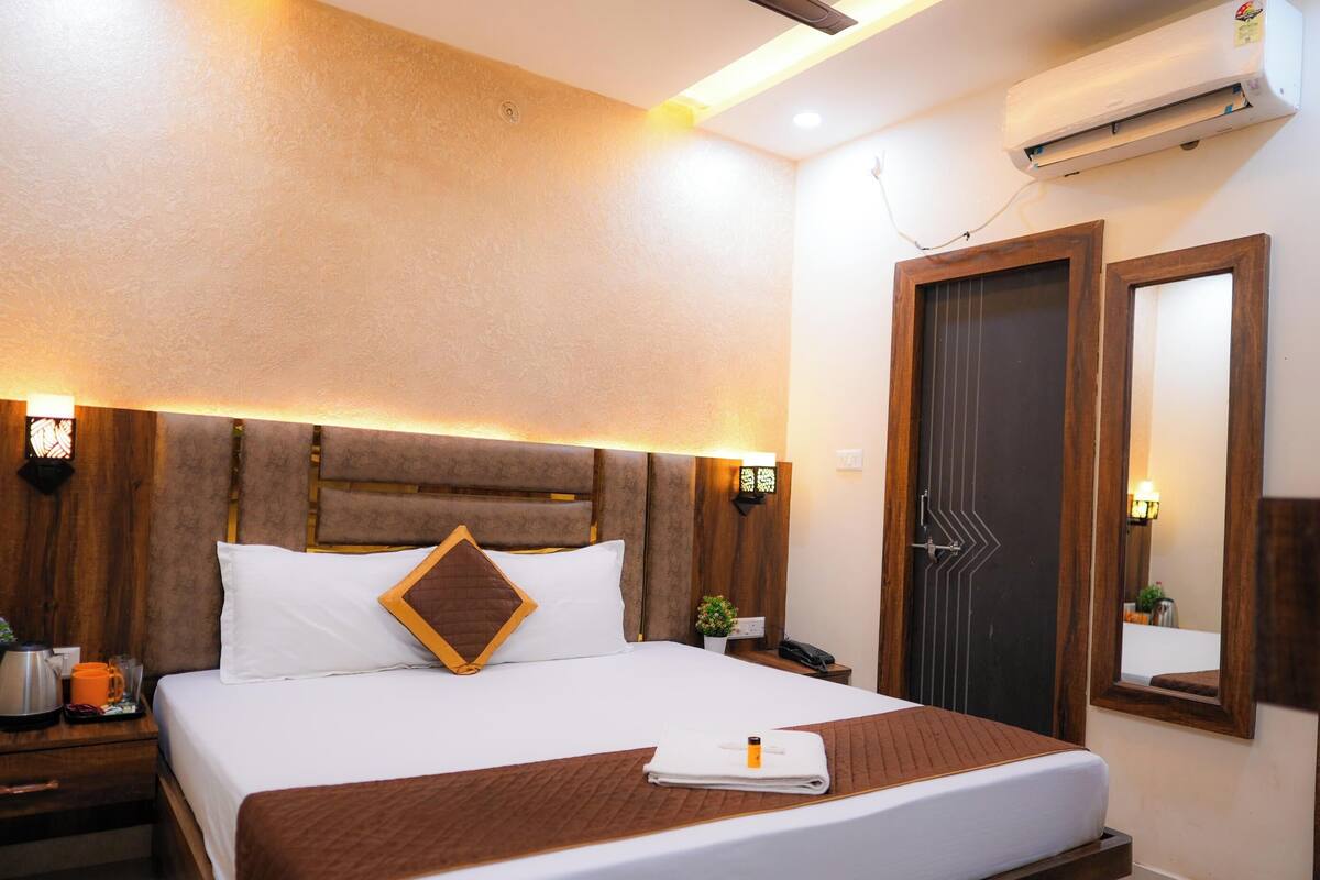 A hotel room with a large bed, wooden headboard, bedside tables, an air conditioning unit, a mirror, and a door with a geometric pattern.