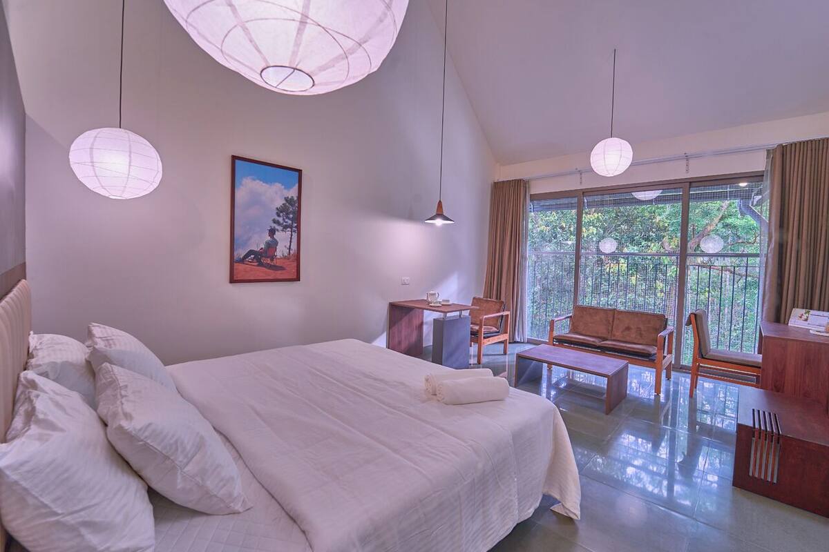 A spacious bedroom with a large bed, a desk, and a chair. There is a balcony with a view of trees.