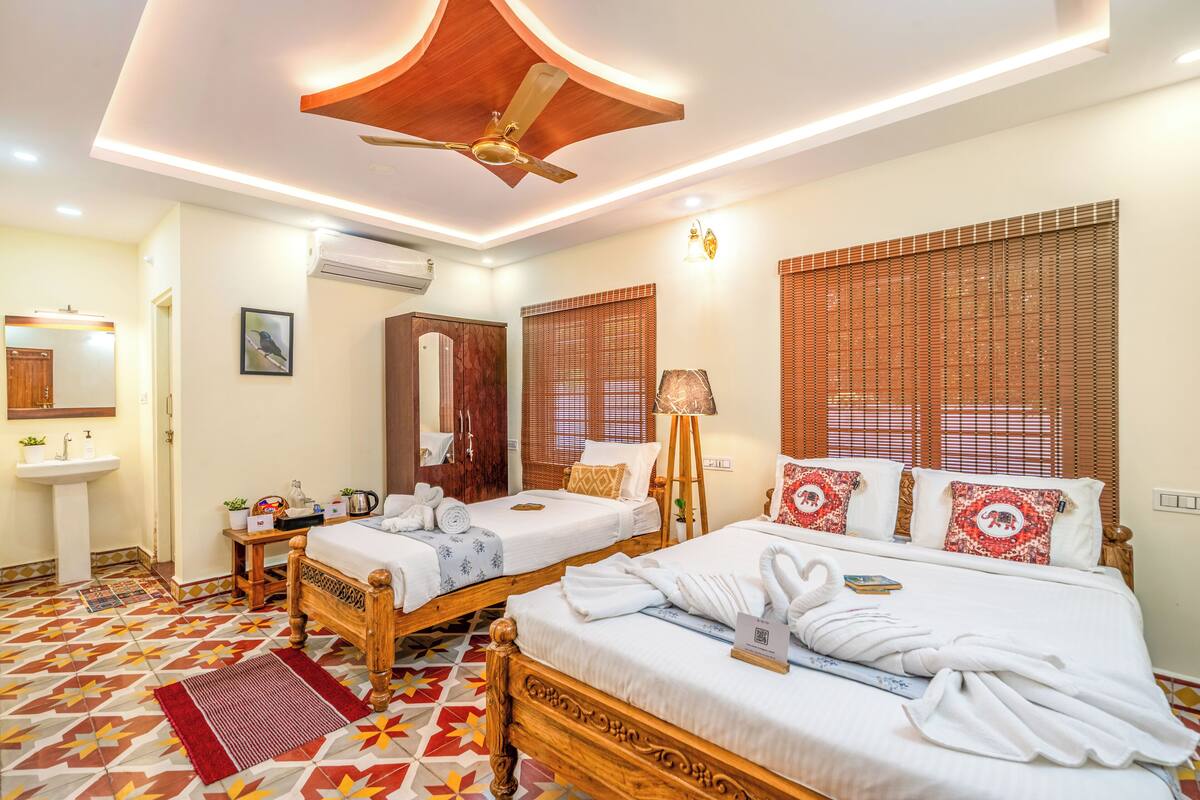 Une chambre d’hôtel avec deux lits en bois, un ventilateur de plafond, une grande fenêtre avec stores, une table de chevet avec une lampe, et une salle de bain avec un lavabo et un miroir.
