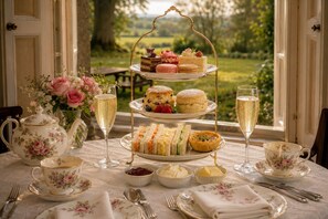 Ein mehrstufiges Teeservice mit Sandwiches, Scones und Gebäck, eine blumengeschmückte Teekanne und Blick auf einen Garten durch ein Fenster.