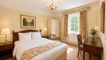 A bedroom with a large bed, two bedside tables with lamps, a desk with a chair, a chandelier, a window with floral curtains, and a painting on the wall.