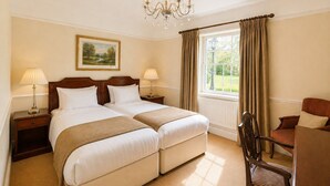A hotel room with two beds, a chandelier, a painting, a window with curtains, and a chair. - Nent Hall Country House Hotel (Alston)
