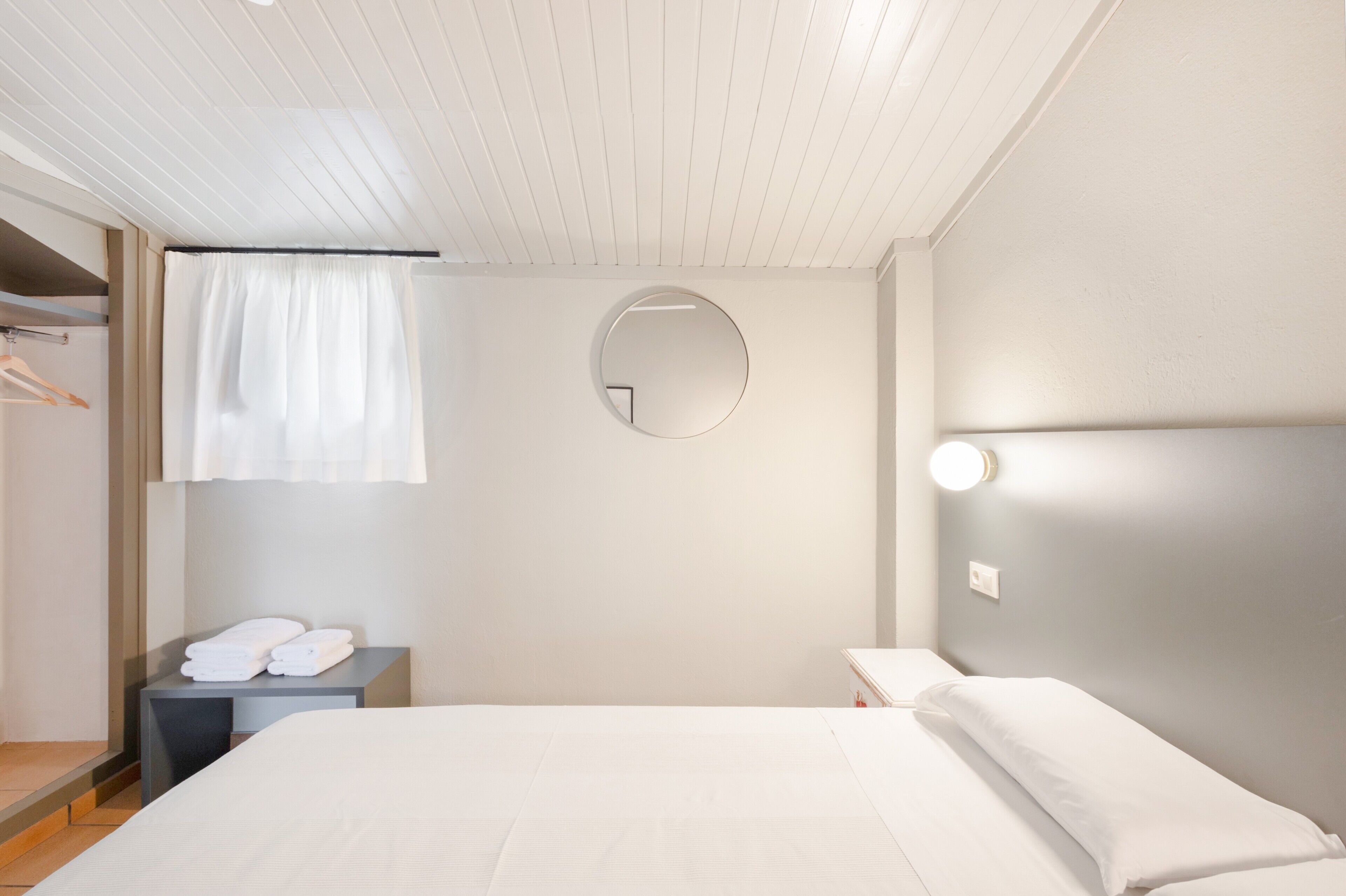 Habitación de hotel con una cama, una mesita de noche con toallas, un espejo redondo en la pared y una ventana con cortinas.