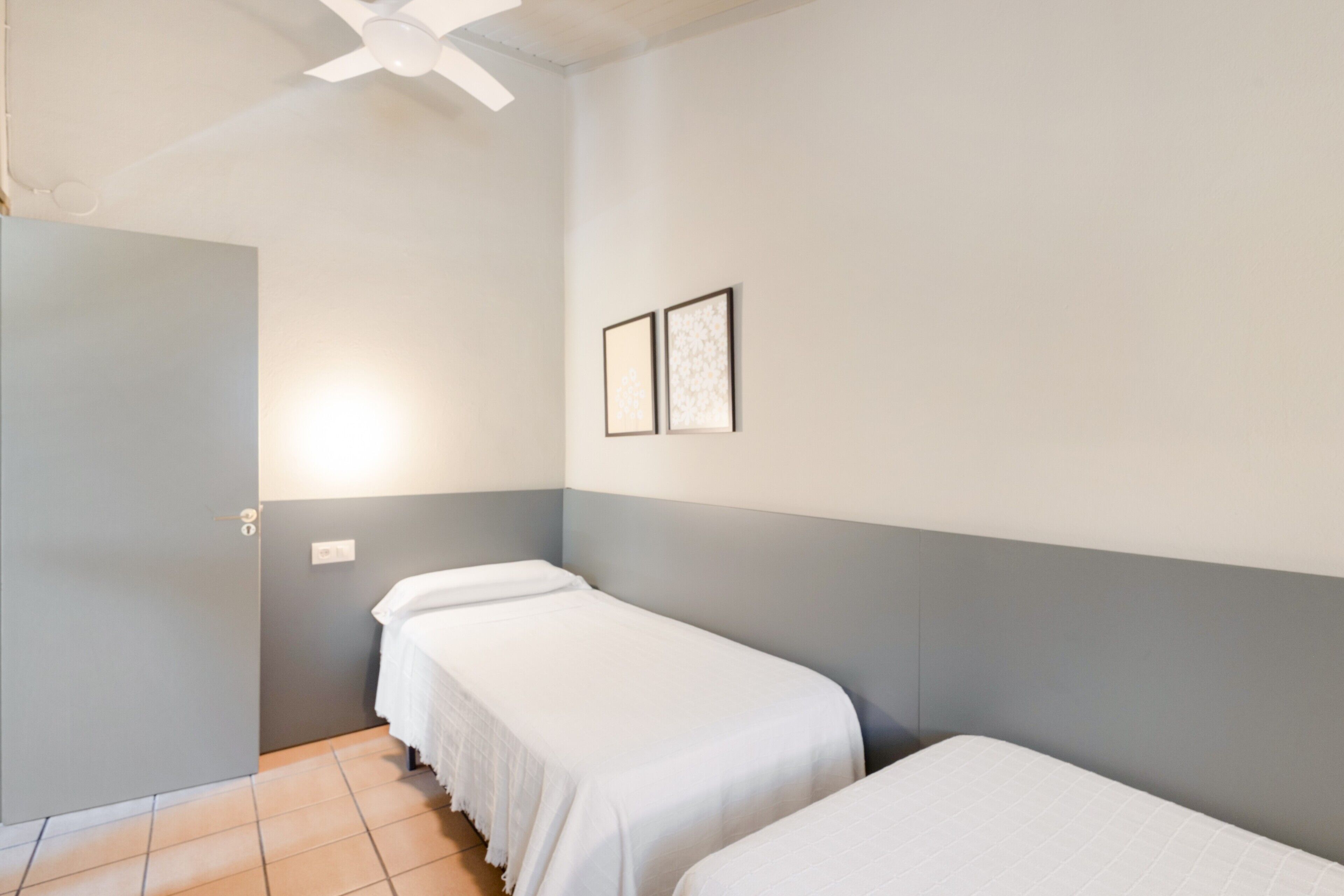 Une chambre avec deux lits simples, un ventilateur de plafond et deux tableaux encadrés accrochés au mur.