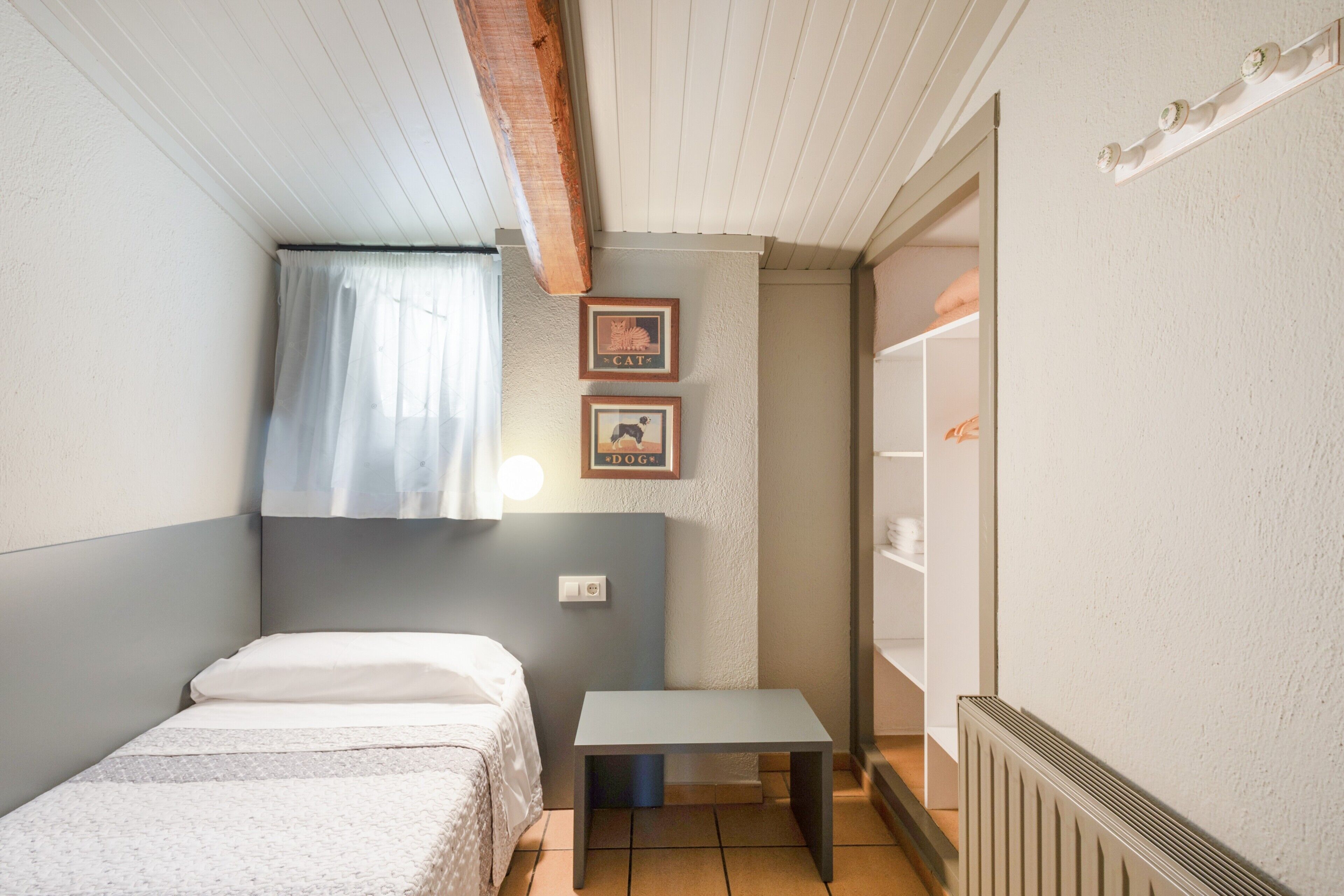 Un dormitorio pequeño y ordenado con una cama individual, una mesita y un ventanal con cortinas.