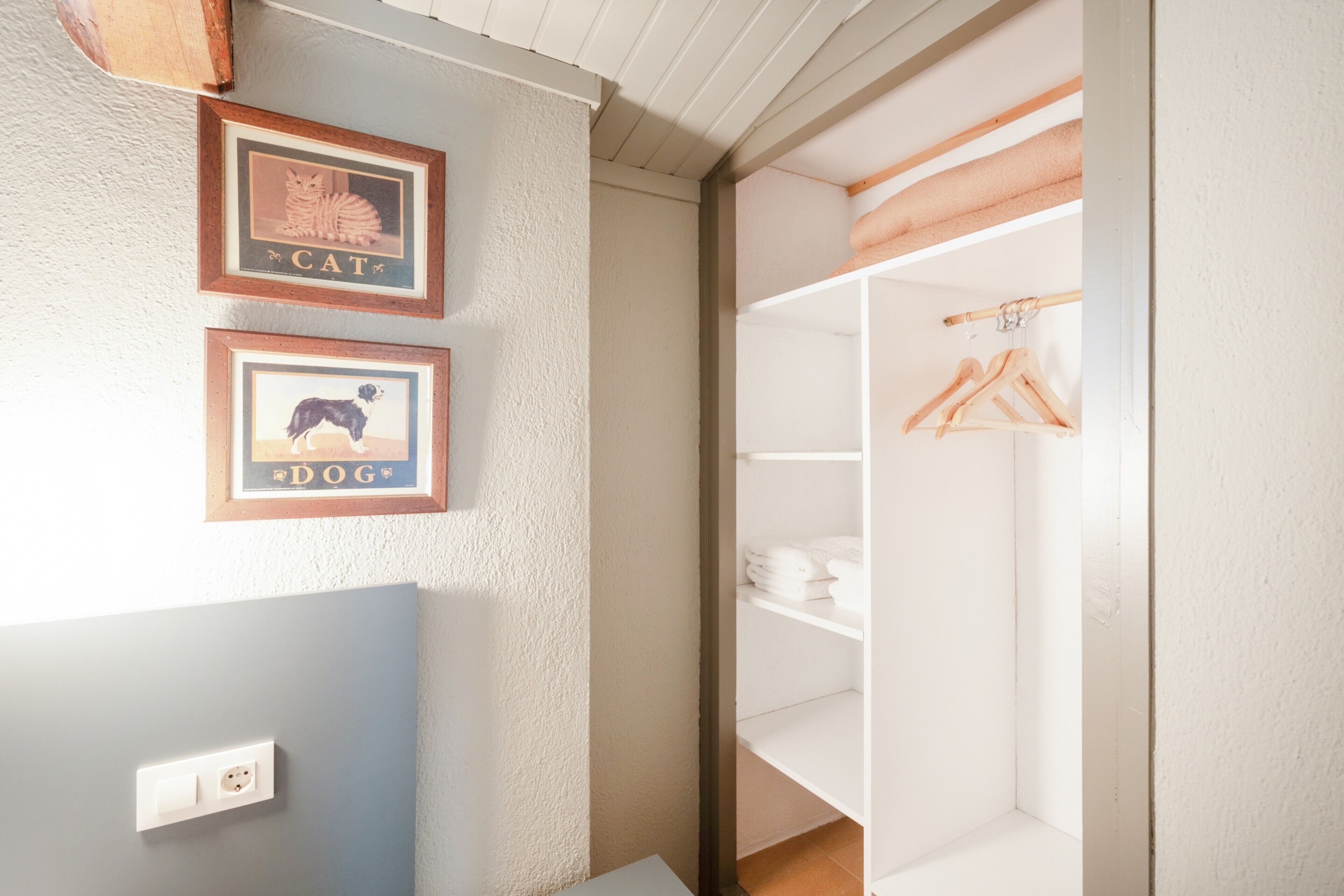 Une pièce avec un mur blanc, un tableau encadré représentant un chat, un tableau encadré représentant un chien, une armoire blanche contenant des vêtements suspendus et une étagère en bois.