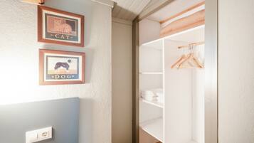 Une pièce avec un mur blanc, un tableau encadré représentant un chat, un tableau encadré représentant un chien, une armoire blanche contenant des vêtements suspendus et une étagère en bois.