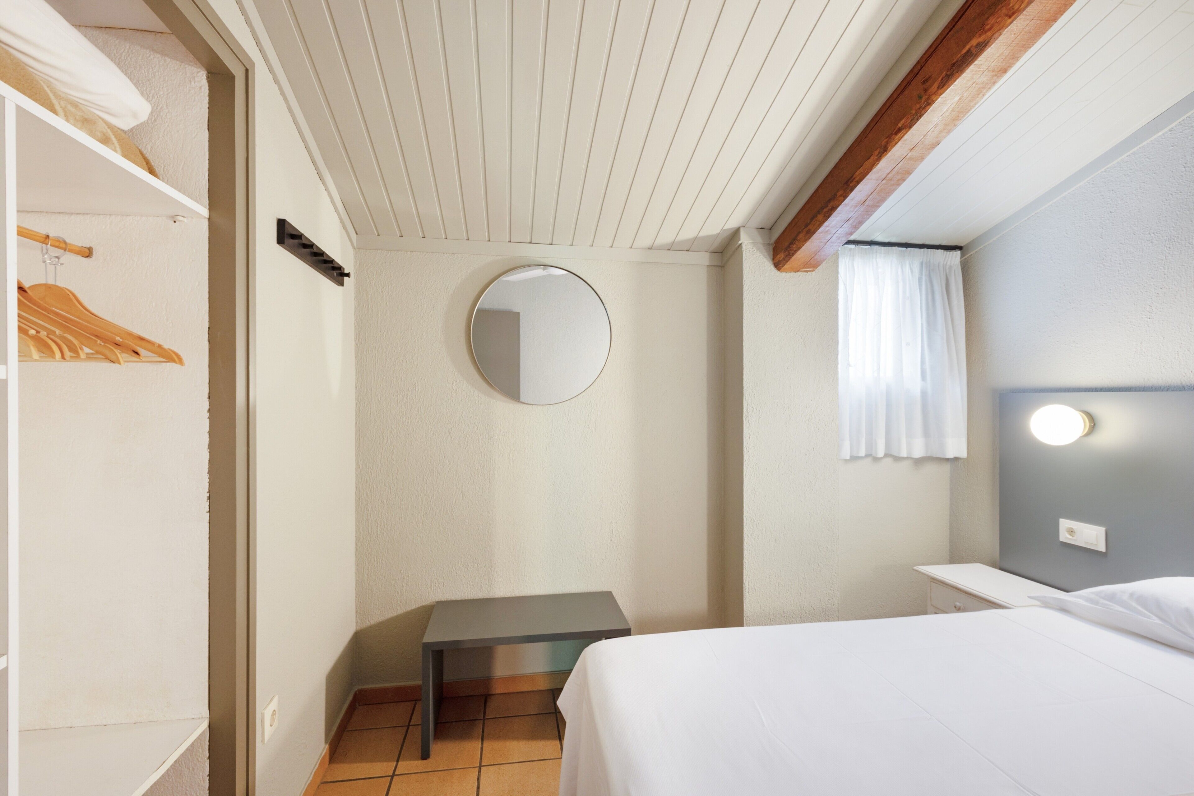 Un dormitorio con cama, mesita de noche, espejo, armario y ventana con cortinas.